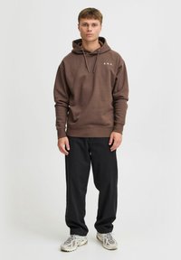 Haut à capuche marron avec le texte blanc "A.R.C.", capuche à cordon de serrage et poignets côtelés, associé à un pantalon noir et des baskets grises. Look simple et décontracté.