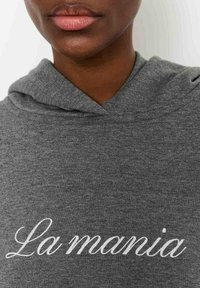 Sivá mikina s jemnou textúrou a nápisom "La mania" bielym kurzívnym písmom zvýrazneným na prednej strane.