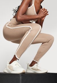 Beige träningsbyxor med vita sidostråkar, kombinerat med vita sportskor, med strukturerad design och rundade sulor.