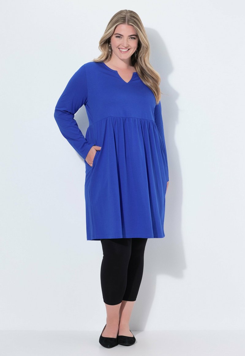 Modré šaty s dlouhým rukávem a výstřihem do V, se rozšířeným pasem a kapsami, spárované s černými legínami a černými balerínami. Hladká textura, ležérní design.