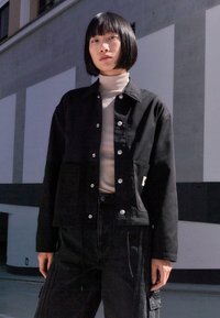 Veste en denim noir avec boutons-pression, col structuré et poche avant. Associée à un pantalon noir taille haute avec des accents verticaux.