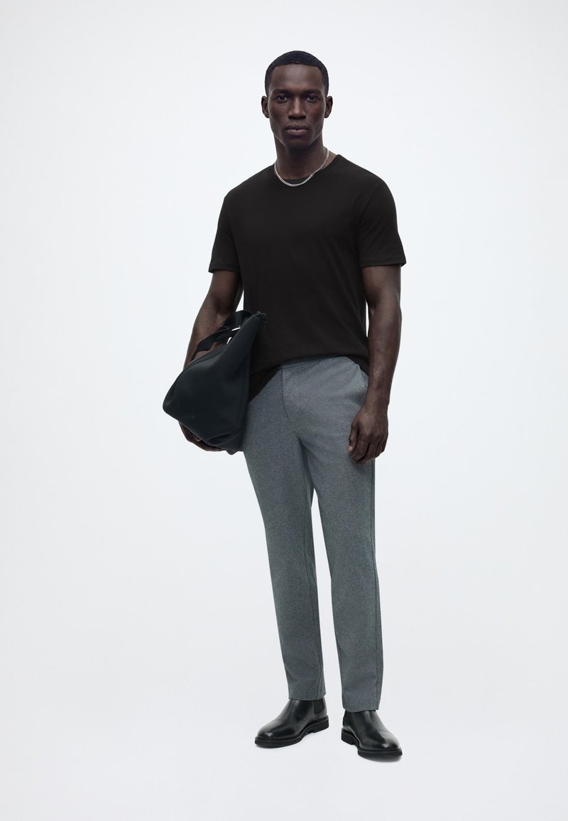 Άνδρας που φορά μαύρο μπλουζάκι, γκρι παντελόνι, μαύρες μπότες, κρατά μια μαύρη τσάντα, στέκεται μπροστά σε απλό φωτεινό φόντο.