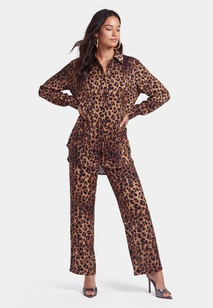 Donna che posa con le mani sui fianchi indossando una camicia a maniche lunghe con stampa leopardata abbinata e pantaloni a gamba larga con sandali con tacco alto.