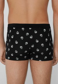 Muž nosící černé boxerky s vzorem malých bílých lebek, zobrazený zezadu od pasu po polovinu stehna.