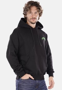 Felpa nera in cotone con tasca a marsupio, cappuccio con cordino e testo del logo verde sul davanti. Texture morbida e vestibilità rilassata.