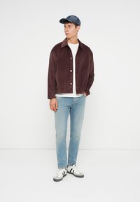 Burgundijas corduroy jaka ar apkakli, balts T-krekls, gaiši zili džinsi un melni un baltie sporta apavi. Modelis valkā džinsa cepuri.