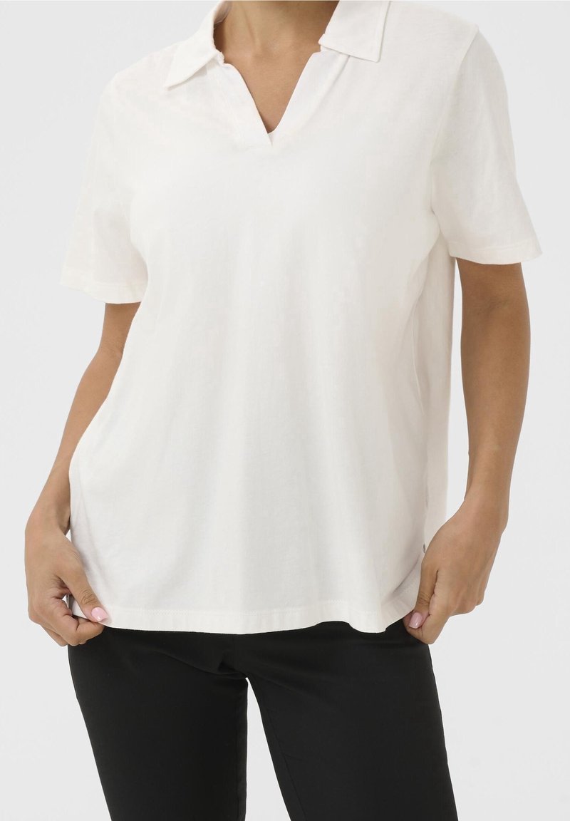 Polo shirt blanc à manches courtes avec un décolleté en V. Tissu doux, coupe décontractée, avec fentes sur les côtés. Associé à un pantalon noir ajusté.