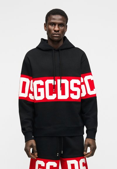 Uomo che indossa una felpa con cappuccio nera e pantaloncini con bande rosse e bianche con il logo GCDS in grassetto sul petto e sulle cosce, in piedi contro uno sfondo semplice.