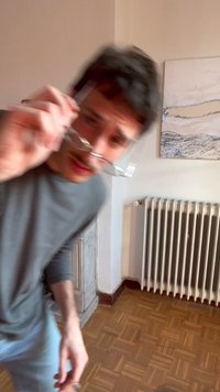 Bărbat tânăr cu cămașă gri ajustând ochelarii în interior, lângă un radiator și un tablou abstract pe perete, ușor neclar.