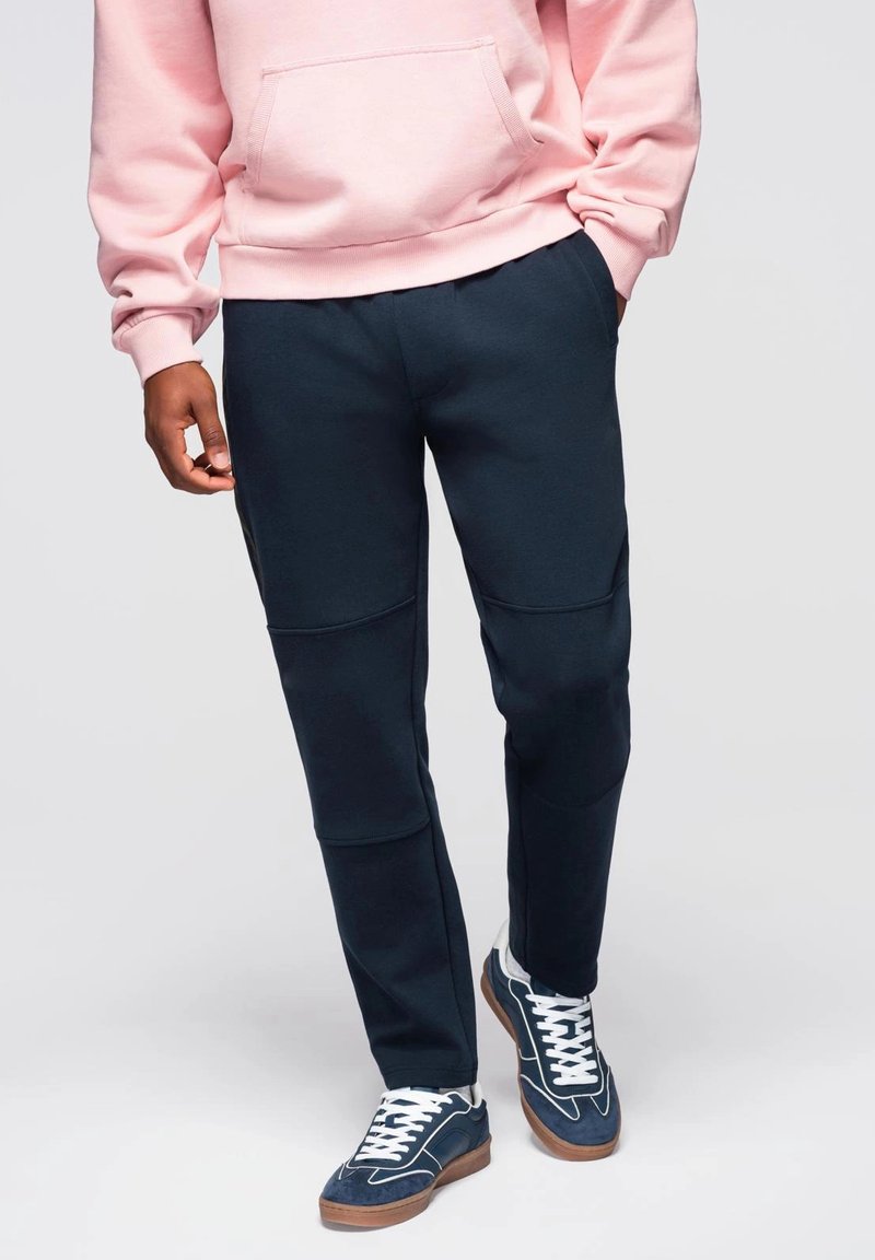 Námořnické jogger kalhoty se bočními panely, kombinované s růžovou mikinou a modrými teniskami. Tkanina se zdá být měkká, s uvolněným střihem a elastickým pasem.