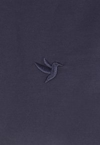Tessuto blu navy con un piccolo design ricamato di un colibrì scuro con le ali spiegate, centrato sulla superficie.