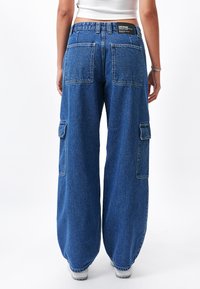 Pantalones cargo de mezclilla anchos y azules con grandes bolsillos traseros, un parche con el logo en la cinturilla y un acabado texturizado.