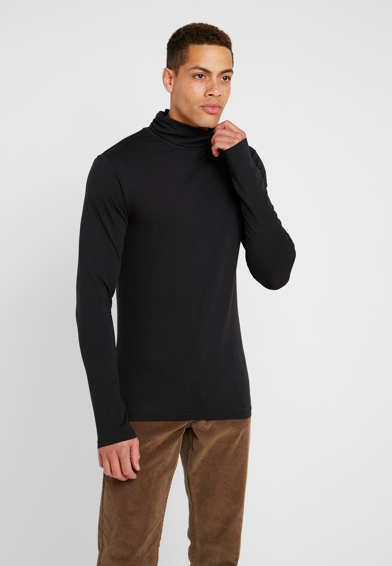 Must särgmoodul, millel on must pika varrukaga kõrge kaelusega turtleneck ja sile kangas, sobib ideaalselt pruunide vooderdatud pükste juurde.