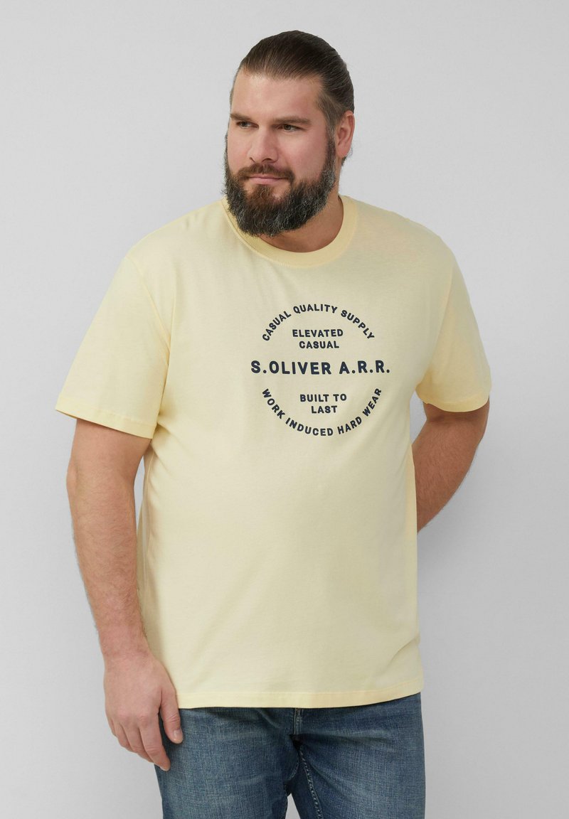 Lichtgele katoenen T-shirt met een ronde hals. Zwarte print in het midden met de merknaam en vetgedrukte teksten. G gedragen met blauwe denim jeans.