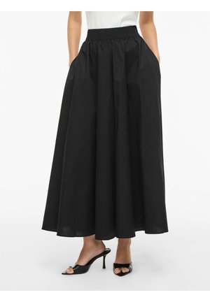 Kvinne iført et svart, hellangt skjørt med høyt liv og lommer, kombinert med svarte åpne hælsandaler og en hvit topp.