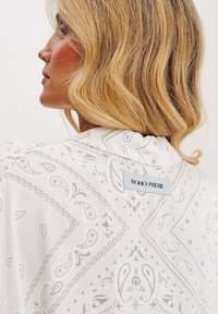 Lehká bílá tkanina s precizními paisley vzory. Má jednoduchý límeček a na zadní straně štítek s nápisem "BOHO PRERIE".
