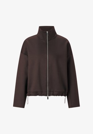 Brūna rāvējslēdzēja jaka no gludas auduma, ar augstu apkakli, brīvu piegriezumu un regulējamu apakšu ar aukliņām.