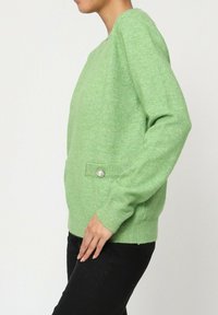 Pull en tricot vert avec des manches longues, un col rond et un tissu texturé. Doté d'une poche latérale avec un détail de bouton décoratif.