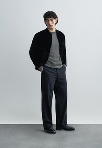 Μαύρο βελούδινο μπουφάν bomber πάνω από γκρι ριγέ πουκάμισο, σε συνδυασμό με ανθρακί παντελόνι και μαύρα παπούτσια. Λείες υφές, εφαρμοστός σχεδιασμός.