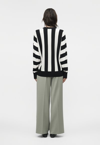 Άτομο που φοράει μαύρο και λευκό πουλόβερ με κάθετες ρίγες και φαρδιά ανοιχτό γκρι παντελόνια, εμφανίζεται από πίσω σε απλό φόντο.