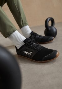 Persoon in zwarte lichtgewicht sportschoenen en witte sokken, zittend bij een zwarte kettlebell op een betonnen vloer.