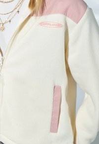 Henkilö pukeutuneena kermaanväriseen fleecetakkiin, jossa on vaaleanpunaiset olkapäälaatat, vaaleanpunaisin reunuksin varustettu tasku ja rinnassa "OFFLINE"-merkki.