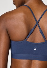 Μπλε αθλητικό σουτιέν σε απόχρωση navy με ριμπ υφή, τιράντες που διασταυρώνονται στην πλάτη, ρυθμιζόμενα στοιχεία και μικρό λευκό λογότυπο στη λωρίδα της πλάτης.