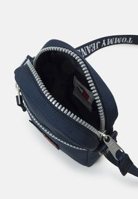 Borsa blu navy con zip realizzata in tessuto durevole. Presenta una zip argentata e una tracolla in tessuto con il testo "Tommy Jeans". Interno foderato.