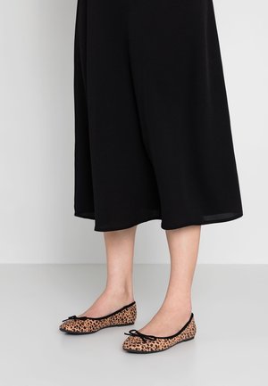 Mujer que lleva una falda midi negra y zapatos de ballet con estampado de leopardo y pequeños lazos negros, de pie sobre un suelo gris claro y fondo blanco.