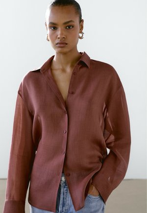 Chemise à manches longues de couleur bordeaux en tissu léger, dotée d'une patte de boutonnage et d'une coupe décontractée avec des fentes latérales, associée à un jean bleu clair.