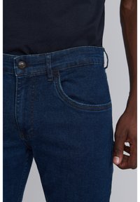 Mörkblå denimjeans med en figursydd passform, fem fickor och knappstängning i midjan. Slät textur och minimala söm detaljer.