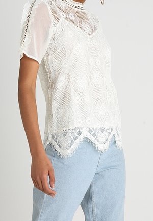 Femme portant un chemisier blanc en dentelle à manches courtes avec des motifs floraux et un ourlet festonné, associé à un jean bleu clair.