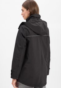 Frau, die eine schwarze wasserdichte Jacke mit Kapuze und reflektierendem Streifen auf dem oberen Rücken trägt, leicht zur Seite geneigt vor einem weißen Hintergrund.