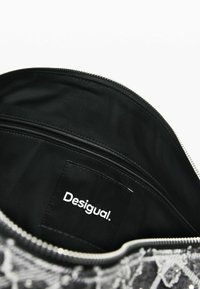 Wnętrze czarnej torby na zamek z kieszenią na zamek; posiada materiałową metkę z napisem "Desigual" na gładkiej, fakturowanej powierzchni.