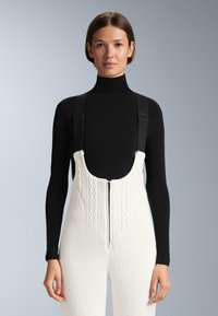 Nő fekete garbót és fehér, texturált kötött kantáros nadrágot visel, vállpántokkal és elöl cipzárral, egyszínű háttér előtt áll.