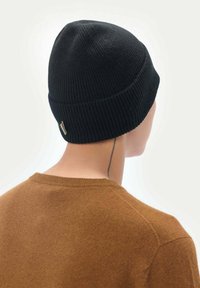 Svart stickad mössa med ribbat mönster och en liten metallmärke, bärs av en person med en brun tröja, visar bakifrån.