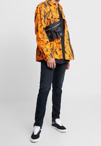 Oranžová košile s černým vzorem větví, černá crossbody taška, tmavé džíny a černé tenisky s bílými podrážkami. Košile má volnější střih.