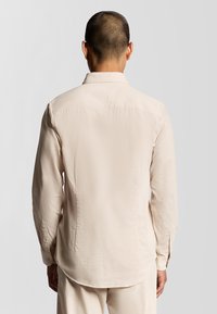 Lyse beige langærmet skjorte lavet af glat stof, med en standardkrave, knapmanchetter og en buet kant bagpå.