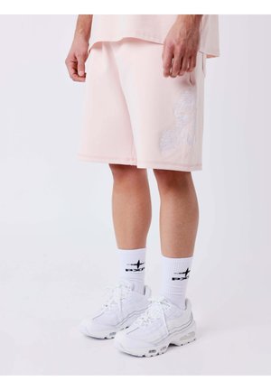 Uomo che indossa pantaloncini rosa chiaro con ricamo floreale, calzini bianchi con marchio e sneakers bianche su uno sfondo semplice.