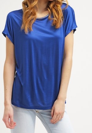 Femme portant un haut bleu ample à manches courtes et un jean bleu clair, debout avec une main près de la hanche et l'autre pendant.
