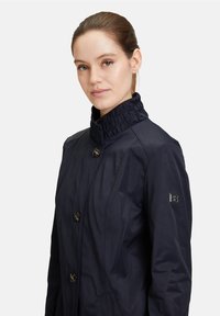 Veste bleu marine avec un col haut, dotée de deux grands boutons et de poches latérales. Le tissu présente une texture lisse, avec un logo sur la manche.