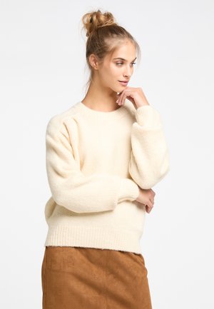 Pull en tricot couleur crème avec un col rond, des épaules tombantes et des poignets côtelés. Porté avec une jupe en daim marron. Texture douce visible.