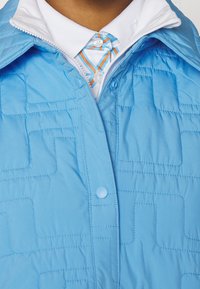 Světle modrá prošívaná bunda s strukturovaným designem a zapínáním na zip. Pod ní je vidět bílá košile s barevnou vzorovanou kravatou.