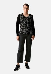 Schwarzes Langarmshirt mit gemusterter Vorderseite, mit Rundhalsausschnitt, kombiniert mit dunkelgrauen geraden Jeans und schwarzen Sportschuhen.