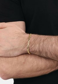 Un uomo con le braccia incrociate che indossa un lucido braccialetto d'oro su un polso peloso, vestito con una camicia nera a maniche corte.