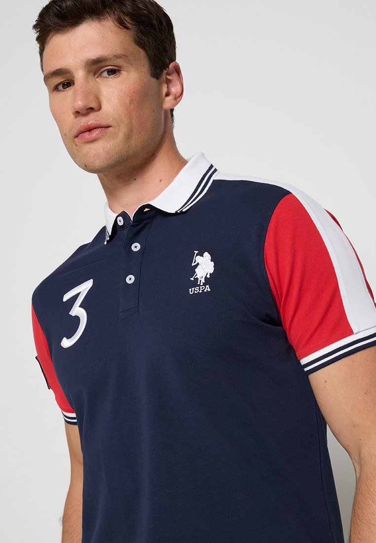 Homme portant un polo bleu marine avec un col blanc, des manches rayées rouges et blanches, le numéro 3 sur la poitrine et le logo USPA sur le côté gauche.