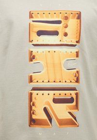 Κείμενο NIKE με τετράγωνα, βιομηχανικού στιλ γράμματα που μοιάζουν με μεταλλικά μέρη με βίδες, με ένα μικρό σήμα Nike σε ανοιχτό ύφασμα φόντο.