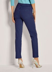 Vrouw in slim-fit marineblauwe broek en pastelkleurige, gedessineerde hoge hakken, staande tegen een effen lichte achtergrond.