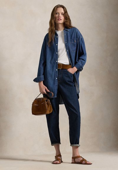 Frau trägt lockeres Jeanshemd über weißem Top, dunkle, hochgekrempelte Jeans, braunen Gürtel, Sandalen, hält eine kleine braune Ledertasche vor einfarbigem Hintergrund.