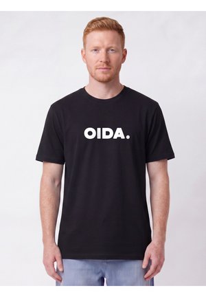 Homme à la peau claire et aux cheveux roux portant un t-shirt noir avec un texte blanc "OIDA.", debout devant un fond clair uni.
